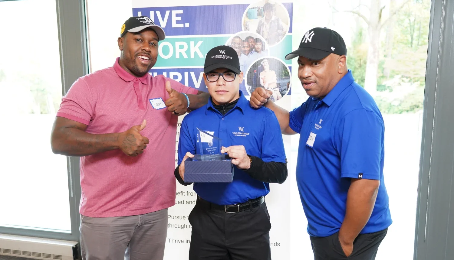 What appears to be three men indoors; center man in blue polo holds a glass trophy in a box, flanked by two thumbs-up teammates by banner.