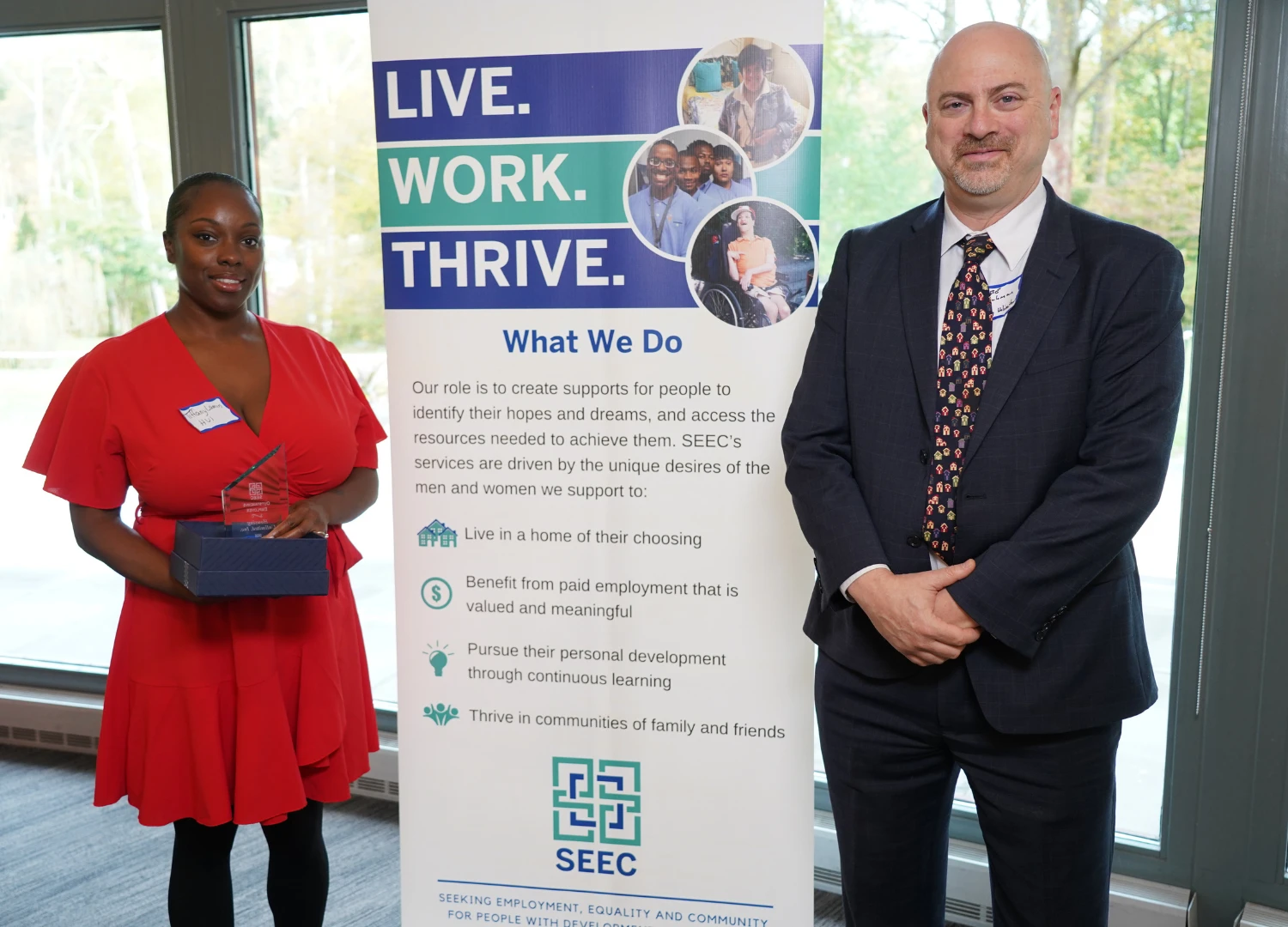 What appears to be a woman in red dress holding a glass award in a blue box, and what appears to be a man in a suit standing beside a SEEC banner.