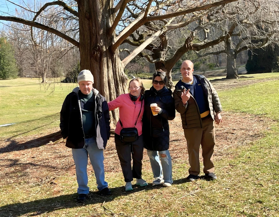 Four adults stand under a bare tree in a sunny park, smiling; one holds a beer while others wear jackets and jeans.
