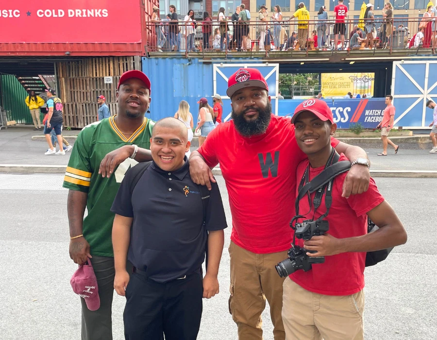 Four friends pose on a busy street festival: green jersey, bald man in black polo, bearded man in red, and a photographer.