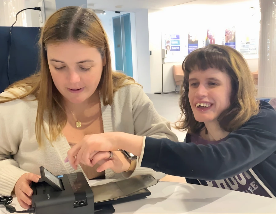 What appears to be two women at a table using a small device; one operates it while the other guides and smiles.