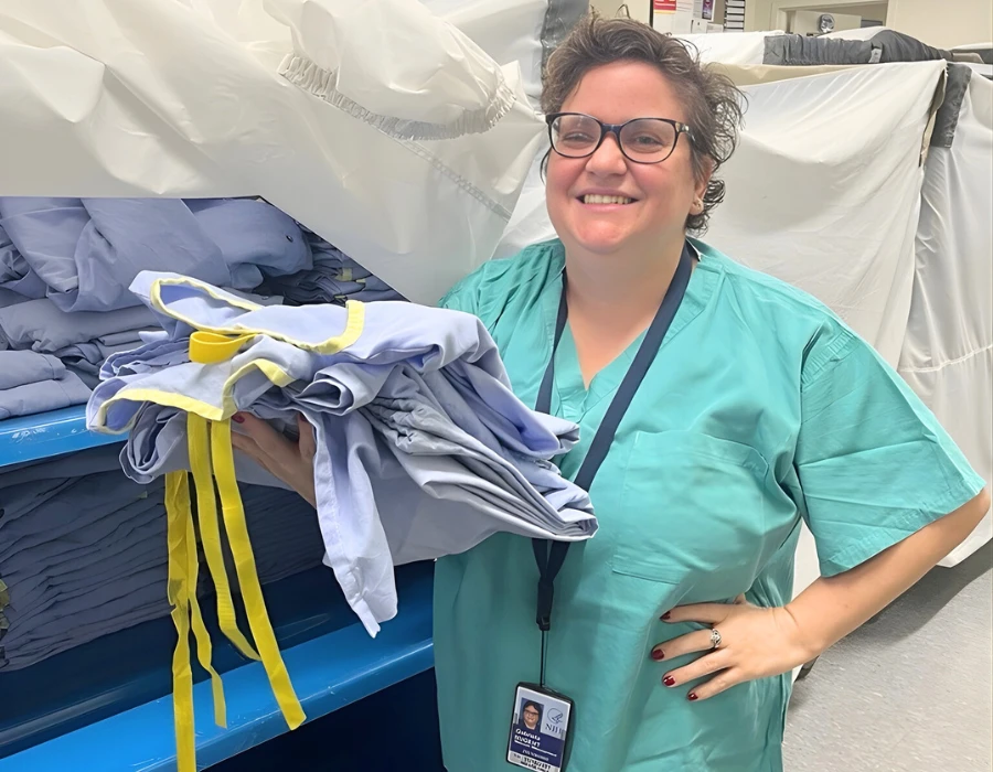 Smiling healthcare worker in teal scrubs holds a stack of blue gowns tied with yellow ribbons in a storage area.