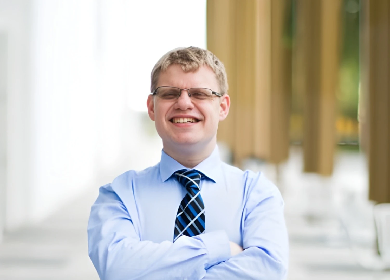 What appears to be a man smiling in blue shirt and striped tie stands with arms crossed, wearing glasses outdoors against blurred column backdrop.