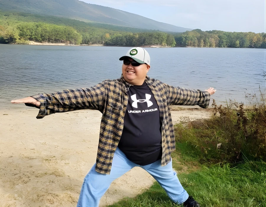 What appears to be a cheerful man in plaid shirt and cap stretches arms wide on a sandy lakeside, with trees and distant mountains behind.
