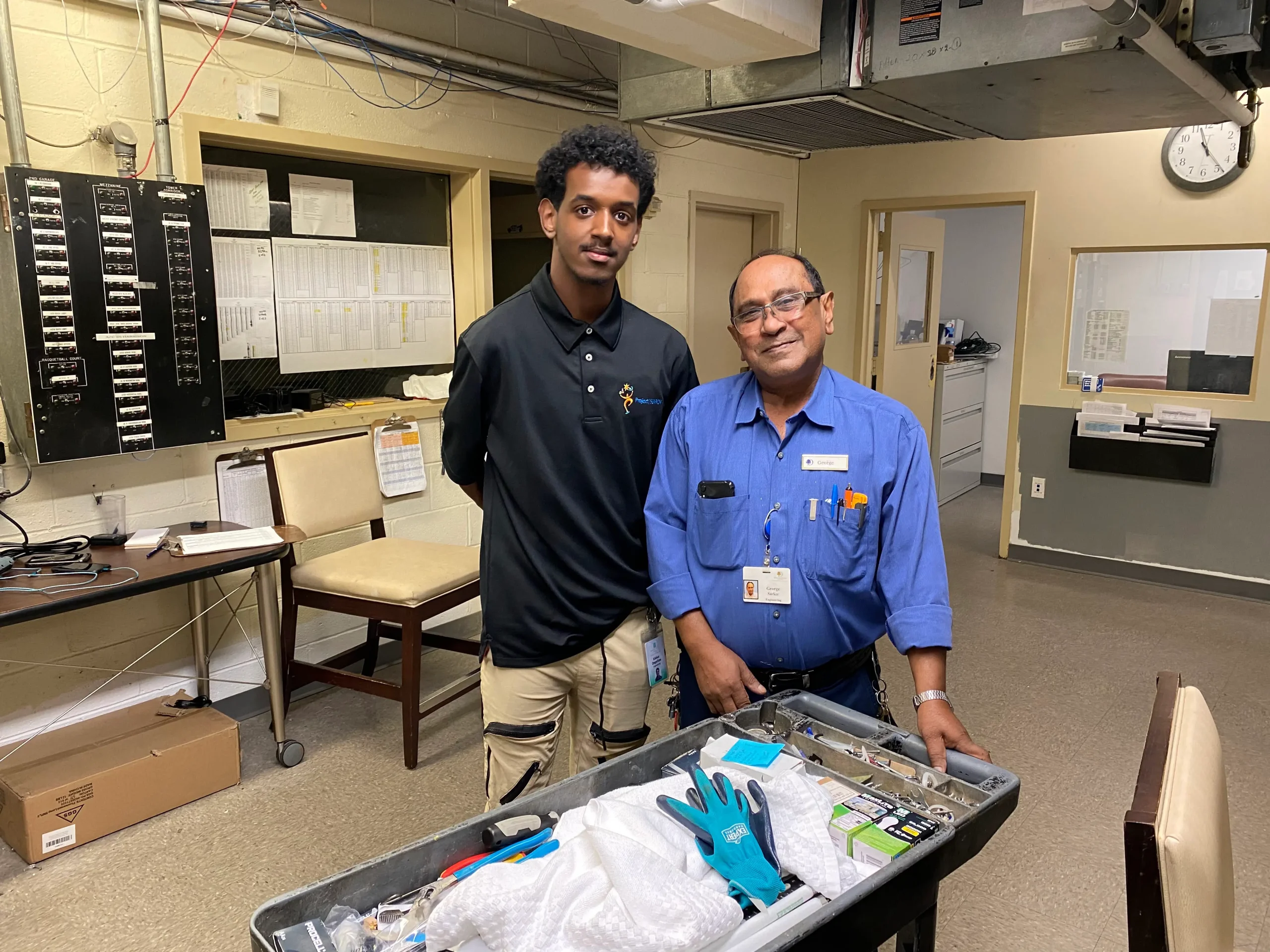 What appear to be two men standing in a maintenance room beside a tool cart; one wears a black polo and khaki pants, the other a blue shirt with an ID badge.