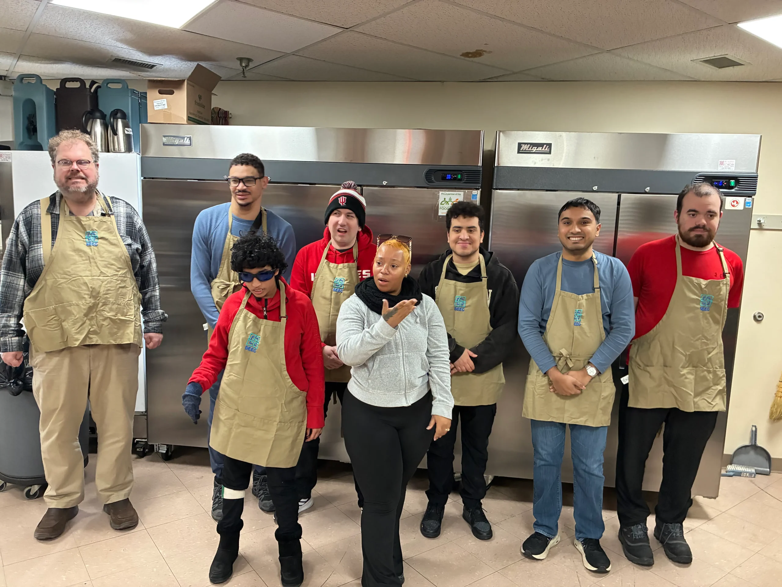 Group of volunteers in beige aprons pose for a group photo in a kitchen with large stainless refrigerators behind them.